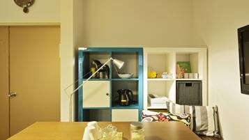 A room with a wooden table, a blue shelving unit with kitchen items, a clock on the wall, and a television on the right.