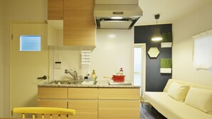 A compact kitchen with wooden cabinets, a built-in oven, and a dining area with a yellow chair. - Konpeito House (Osaka)