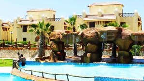 A swimming pool with a dinosaur-themed water feature and a building with a tiled roof in the background. - Marseilia Beach 3 (Al-Hammam)