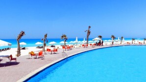 A poolside area with lounge chairs and umbrellas, a clear blue sky, and a beach in the background. - Marseilia Beach 3 (Al-Hammam)