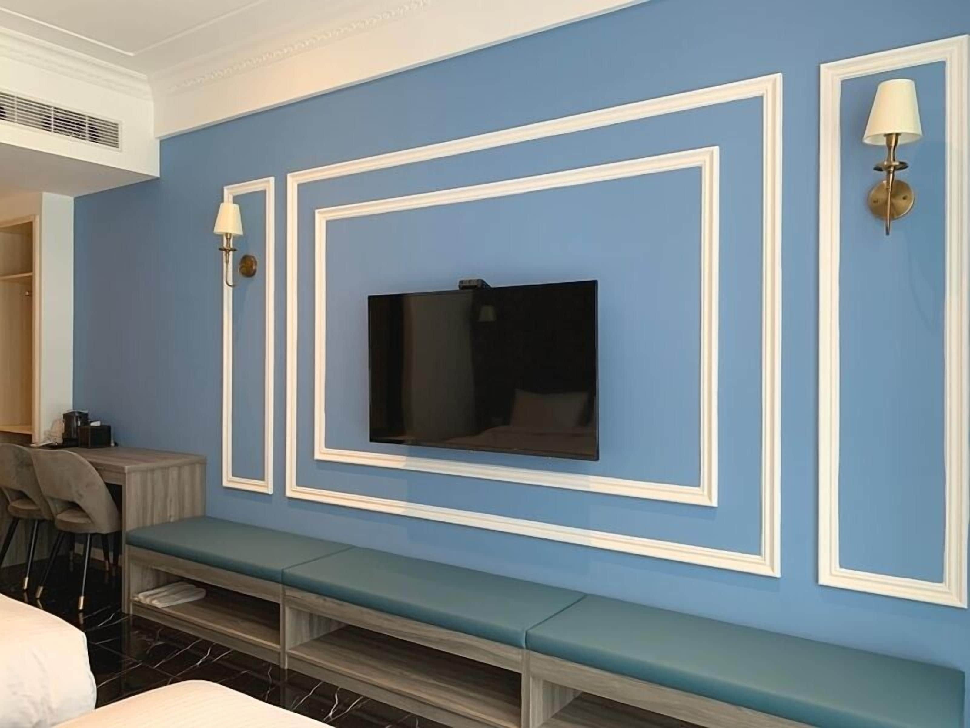 A hotel room with a large flat-screen TV mounted on a blue wall, a bench, and a desk with a chair.