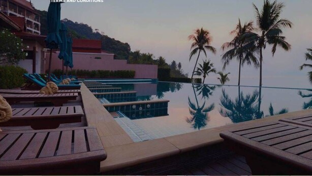 Zona de piscina con sillones de descanso y vistas a palmeras y un edificio al fondo.