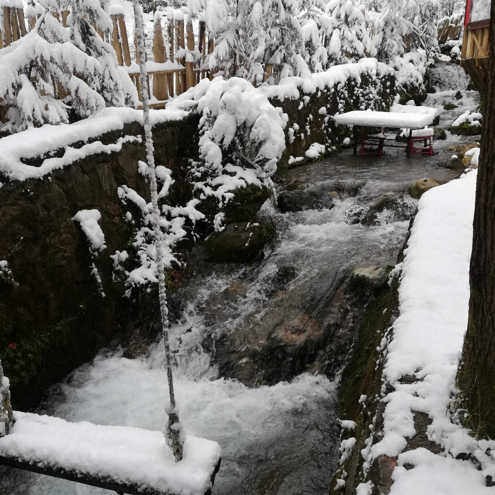 Zona exterior cubierta de nieve con un pequeño arroyo, bancos de madera y una valla de madera.