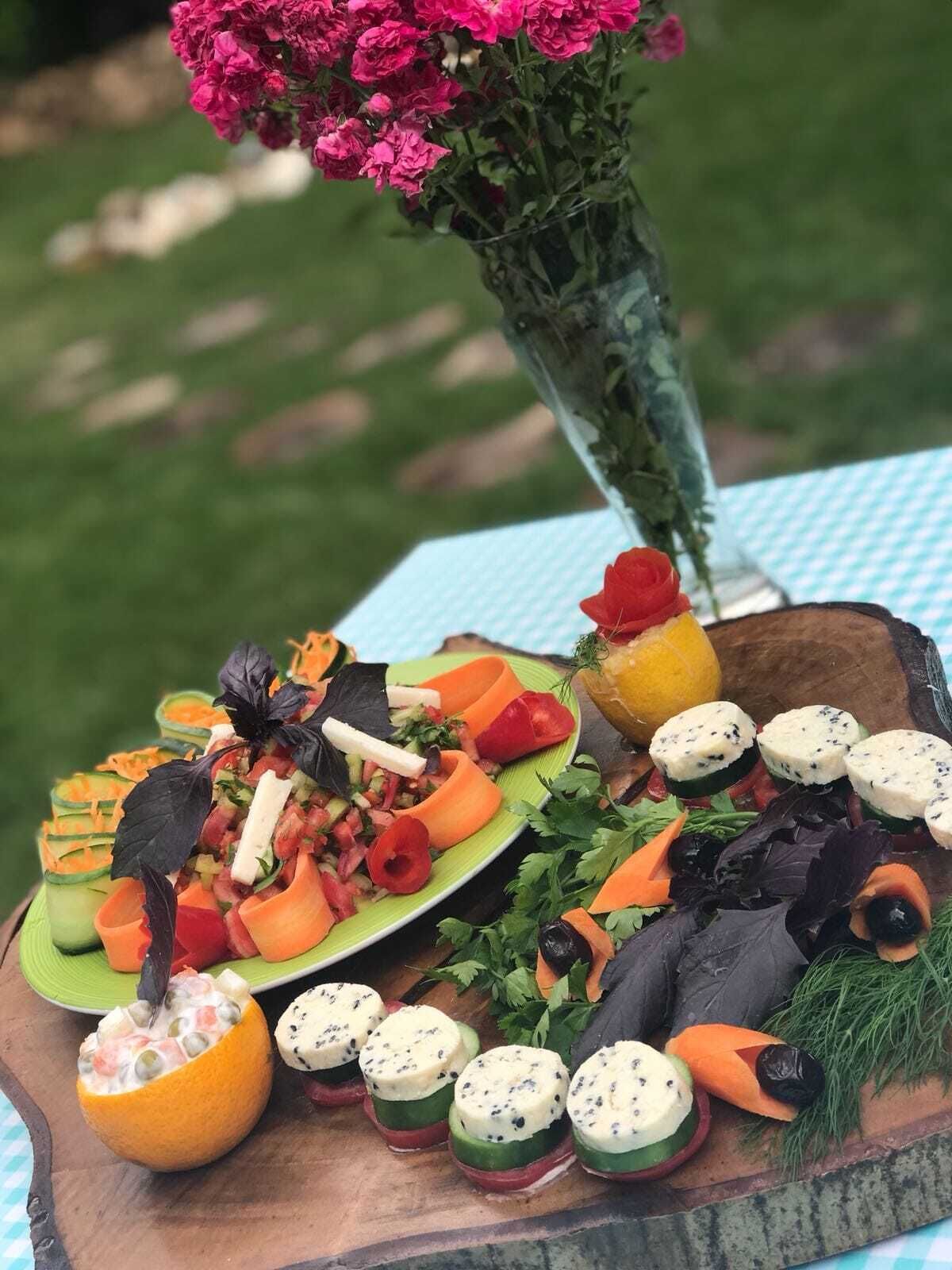 Uma tábua de madeira com diversos alimentos, incluindo uma salada, cubos de queijo e um arranjo floral decorativo.