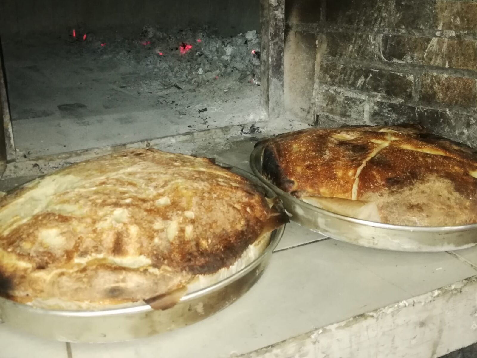 Twee ronde metalen dienbladen met gebak, mogelijk brood, op een witte ondergrond in de buurt van een bakstenen muur.