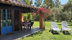 Une maison avec une porte bleue, une terrasse avec une table et des chaises, et deux fauteuils de jardin à rayures dans un jardin.