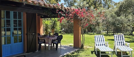 A house with a blue door, a patio with a table and chairs, and two striped lounge chairs in a garden.