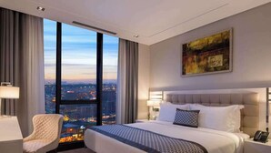A hotel room with a large bed, a chair, a desk, and a view of the cityscape through large windows. - La Verda Suites and Villas (Istanbul)