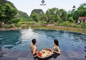 A couple sitting by a pool with a table of food in a lush, green setting. - Maison Que (Nam Hoa Lu)