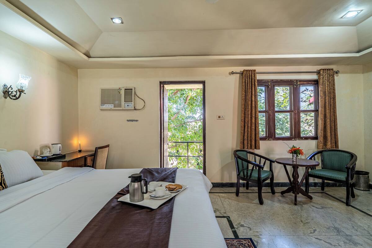 A hotel room with a bed, a desk with a chair, a small table with a vase, and a window with curtains.