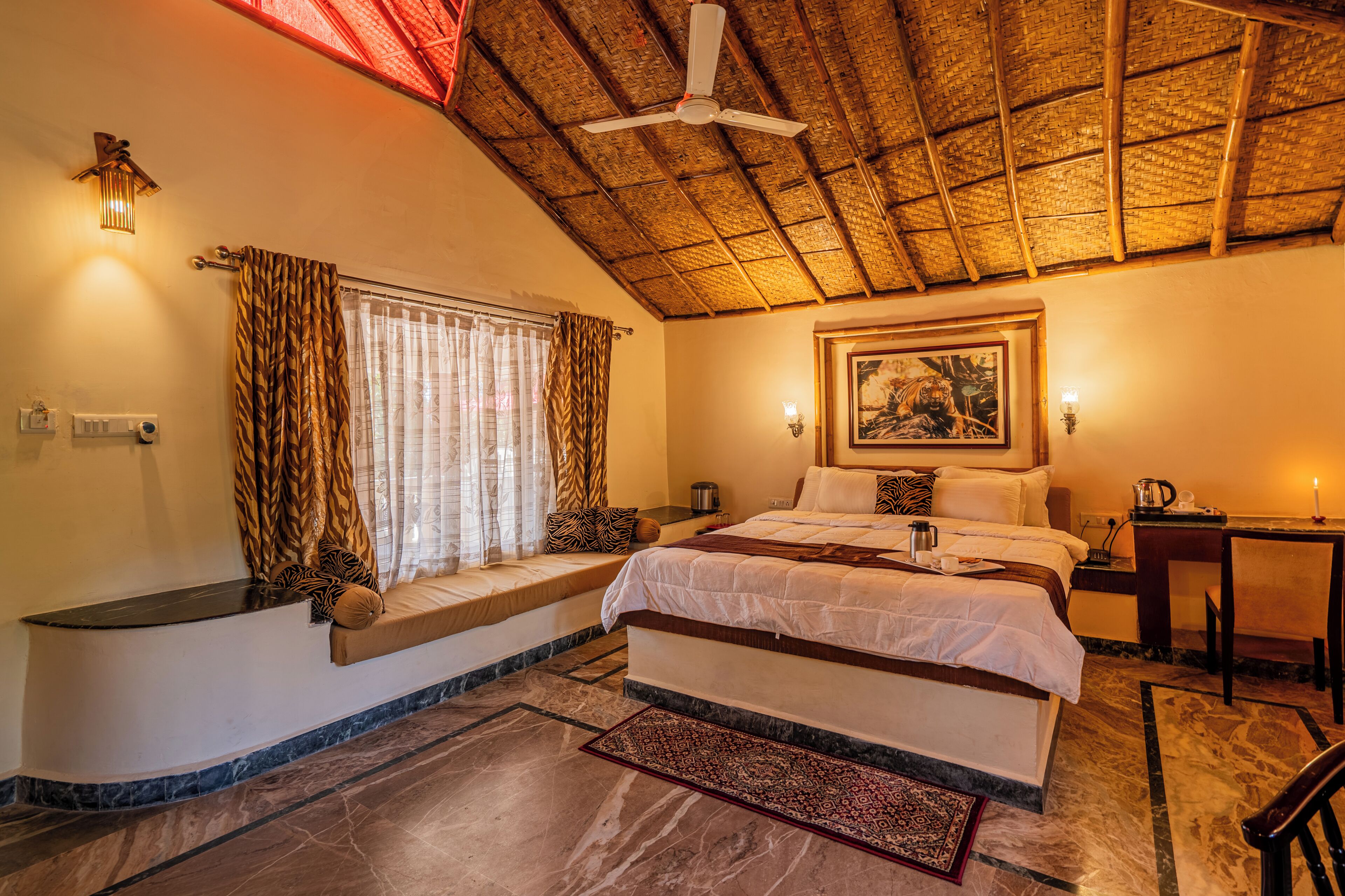 A bedroom with a bed, a window with curtains, a ceiling fan, a painting on the wall, and a small table with a chair.