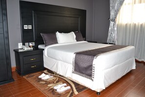 A neatly made bed with a headboard, a bedside table with a phone and a lamp, and a pair of slippers on a rug. - Panone Hotel Boma (Arusha)