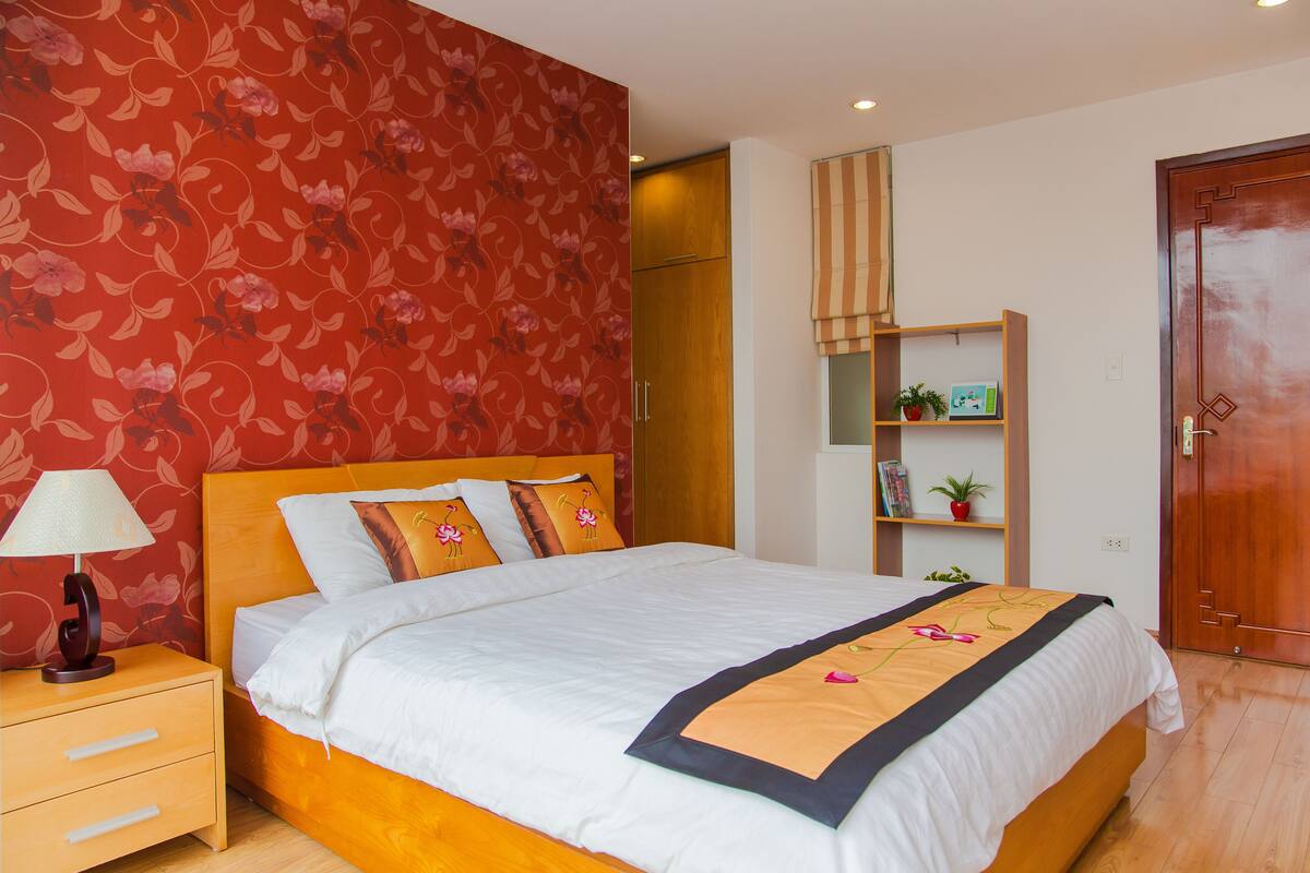 Un lit bien fait, recouvert d’une couvre-lit blanche et beige, une table de chevet en bois avec une lampe, et un papier peint à motifs floraux rouges.