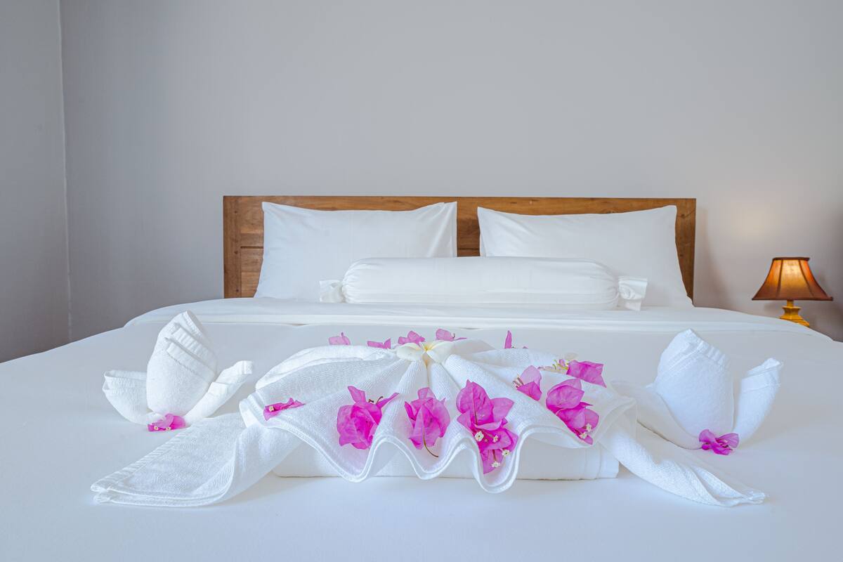 A neatly made bed with white linens and pink flowers, a wooden headboard, and a bedside lamp.