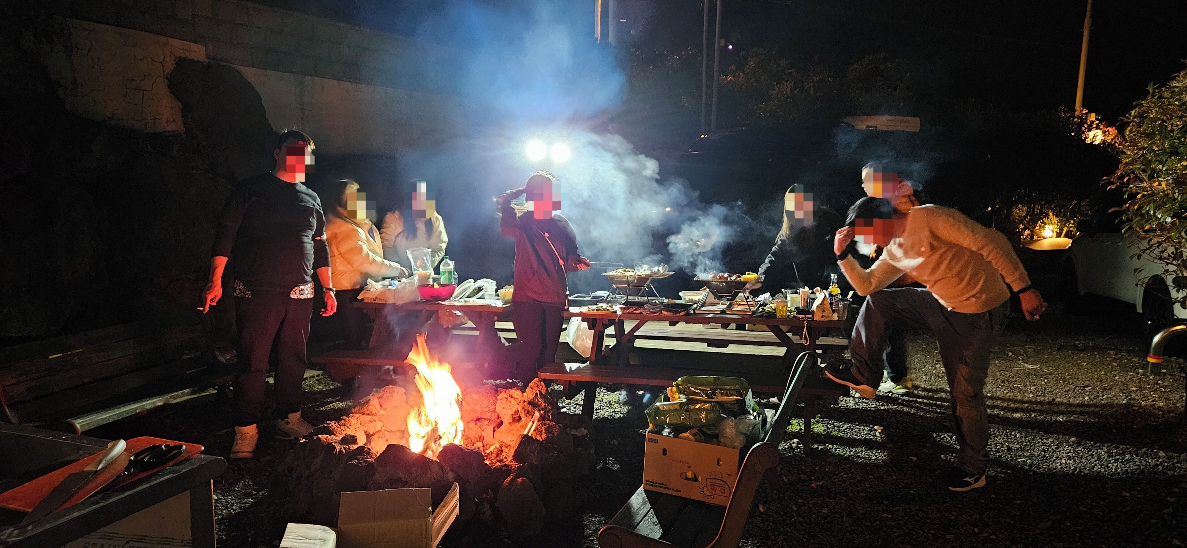 Un groupe de personnes s’est rassemblé autour d’un feu de camp, avec de la nourriture et des boissons, ce qui laisse supposer qu’il s’agit d’un événement social en plein air.