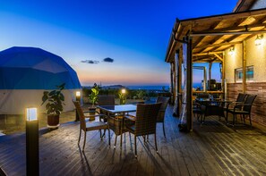 Une terrasse en bois avec des meubles d’extérieur, un espace abrité avec une table et des chaises, et une vue sur l’océan au crépuscule.