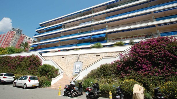 Un hôtel à plusieurs étages, doté de balcons, d’une façade en pierre et d’un parking où sont stationnées des voitures et des scooters.