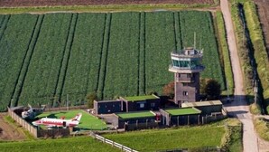 Eine Luftaufnahme einer Farm mit grünen Feldern, einem kleinen Gebäude mit einem Turm und einem abgestellten Flugzeug.