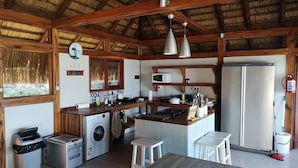 A compact kitchen with wooden beams, a washing machine, a refrigerator, and a dining area with stools. - Aloha 1 (Ponta d'Ouro)
