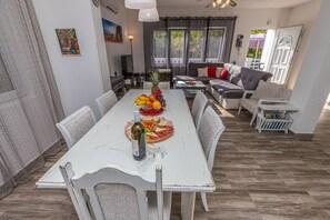 Un coin repas avec une table blanche dressée pour quatre personnes, un panier de fruits et une bouteille de vin. On aperçoit un canapé et une petite table au fond.