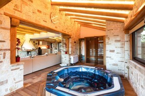 A stone-walled room with a hot tub, wooden flooring, and a kitchen area visible through an open doorway.