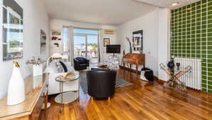 Un salon moderne avec un canapé, des fauteuils, une table basse et un téléviseur. Il y a une fenêtre donnant sur l’extérieur, un radiateur et un mur recouvert de carreaux verts.
