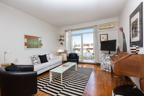 A modern living room with a sofa, coffee table, and a TV. There is a large window with a view of buildings outside.