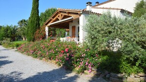 Una casa con porche cubierto, rodeada por un camino de grava y un jardín lleno de flores rosadas.