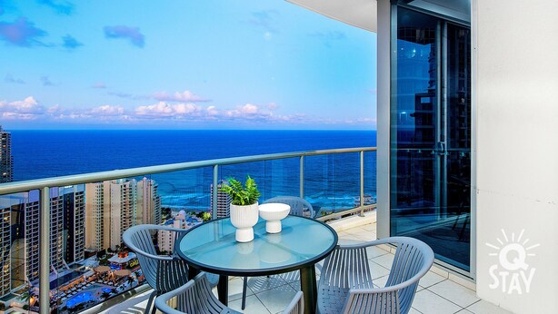 A balcony with a glass table and chairs, overlooking a cityscape and the ocean. - Chevron – 3 Bed Executive Sub Penthouse Ocean (Surfers Paradise)