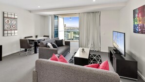 A modern living room with a grey sofa, a dining table, and a flat-screen TV. - H'Residences – 2 Bed City View — Q Stay (Surfers Paradise)