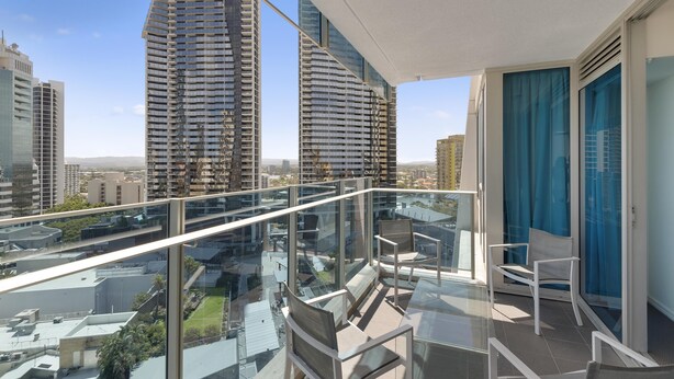 A balcony with a glass railing overlooking a cityscape, featuring modern buildings and a clear sky. - H'Residences – 2 Bed City View — Q Stay (Surfers Paradise)