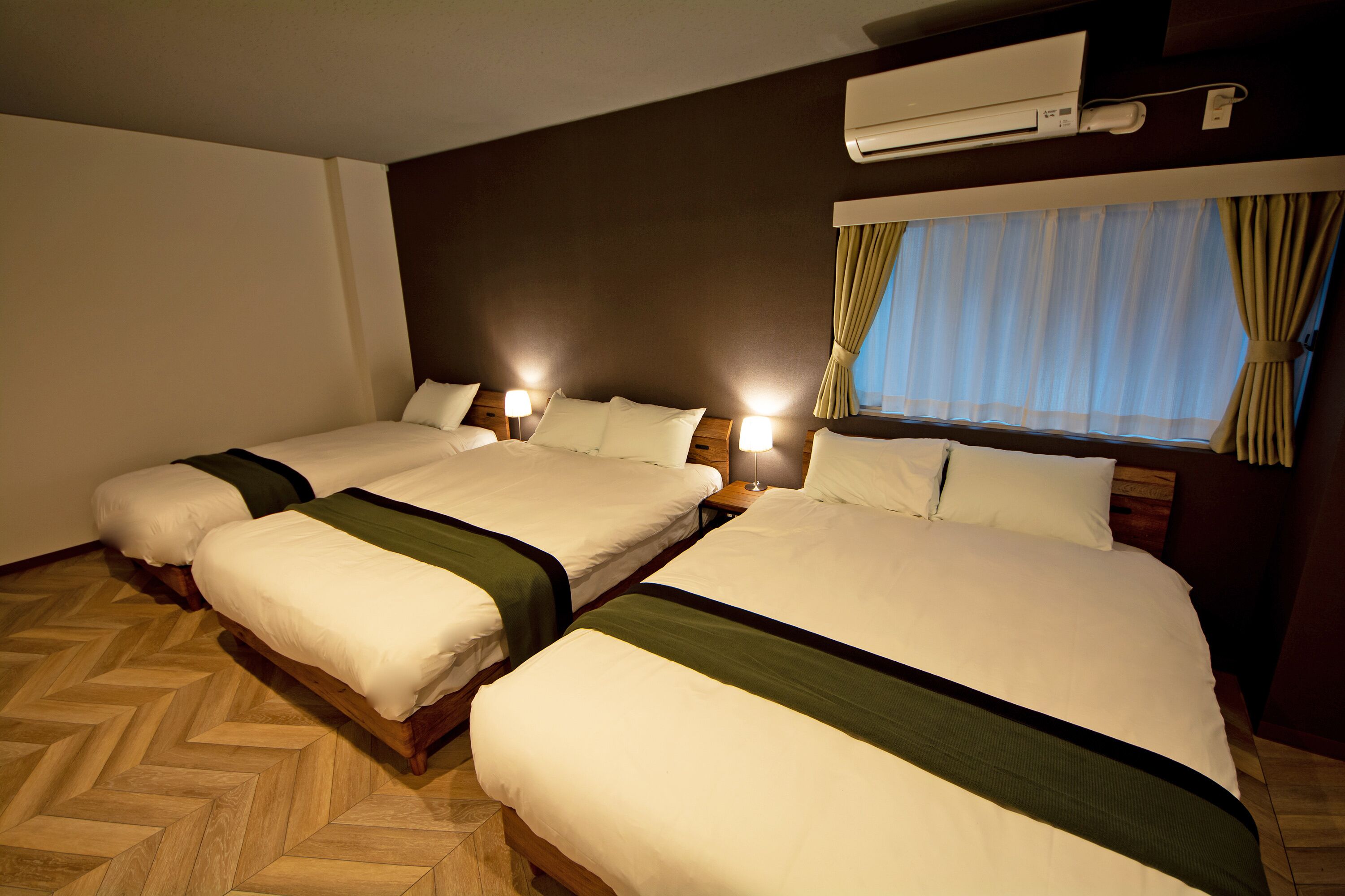 Een hotelkamer met twee bedden, houten vloeren, een raam met gordijnen en een airconditioning.