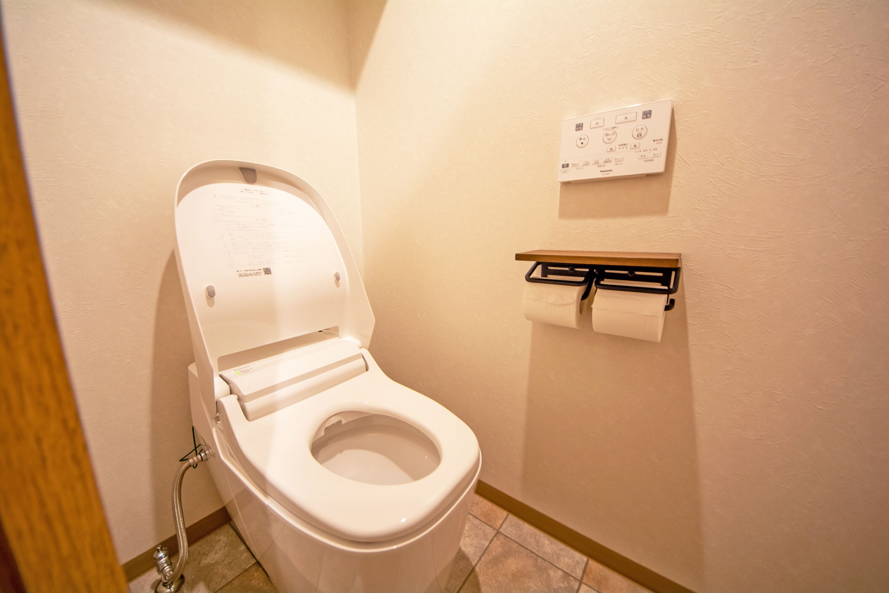 Inodoro blanco con la tapa cerrada, un dispensador de papel higiénico en la pared y el botón de descarga a la izquierda.