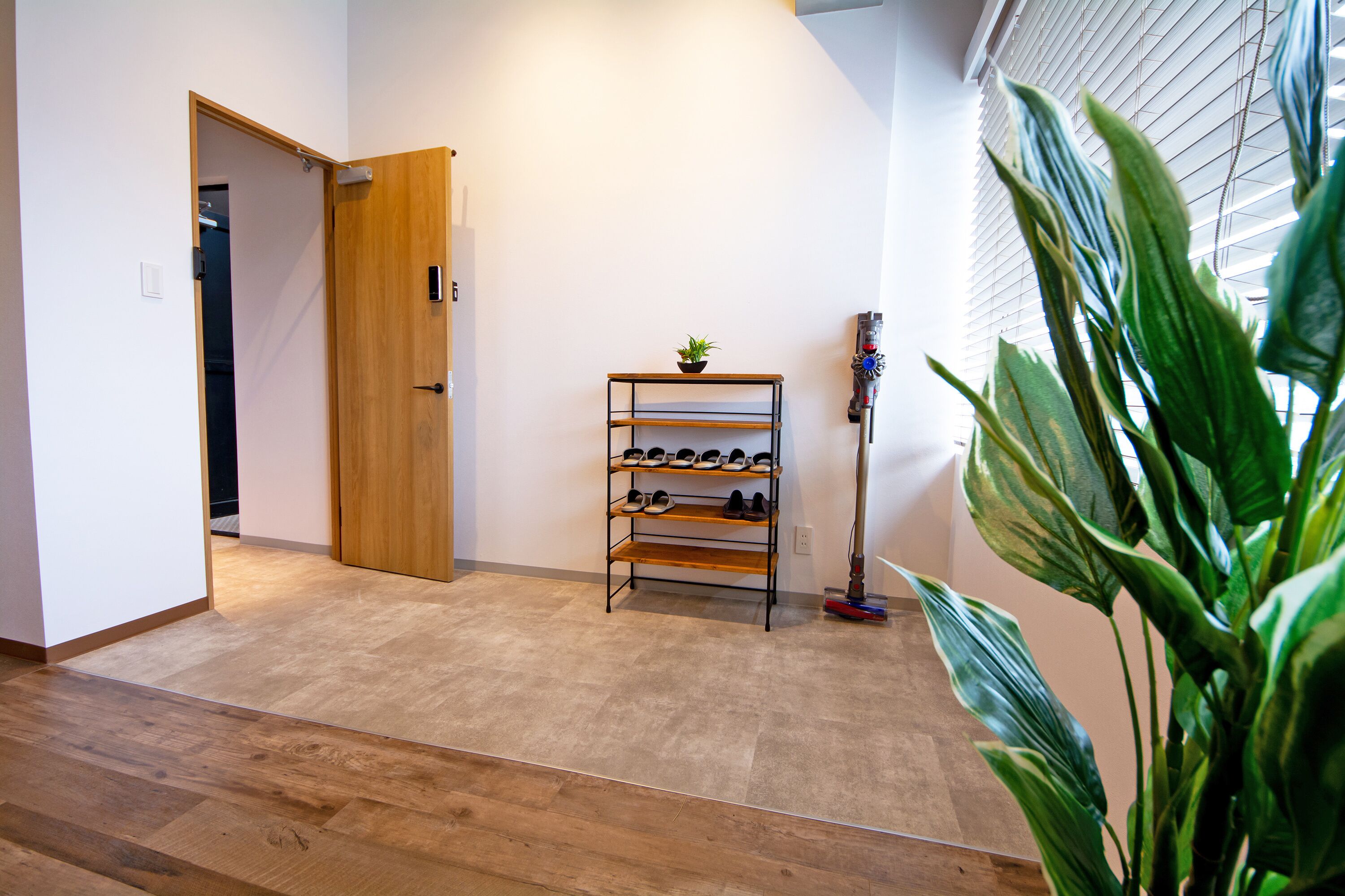 Un couloir avec une porte en bois, un meuble à chaussures, une plante en pot et une fenêtre avec des stores.