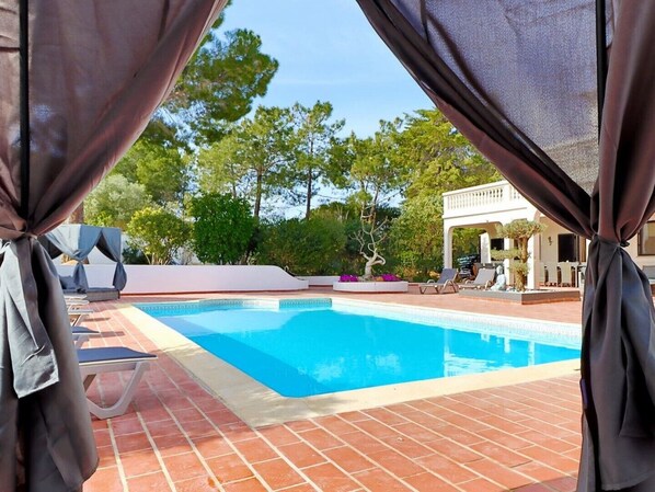 A swimming pool with lounge chairs and a covered area, surrounded by trees and a building in the background.