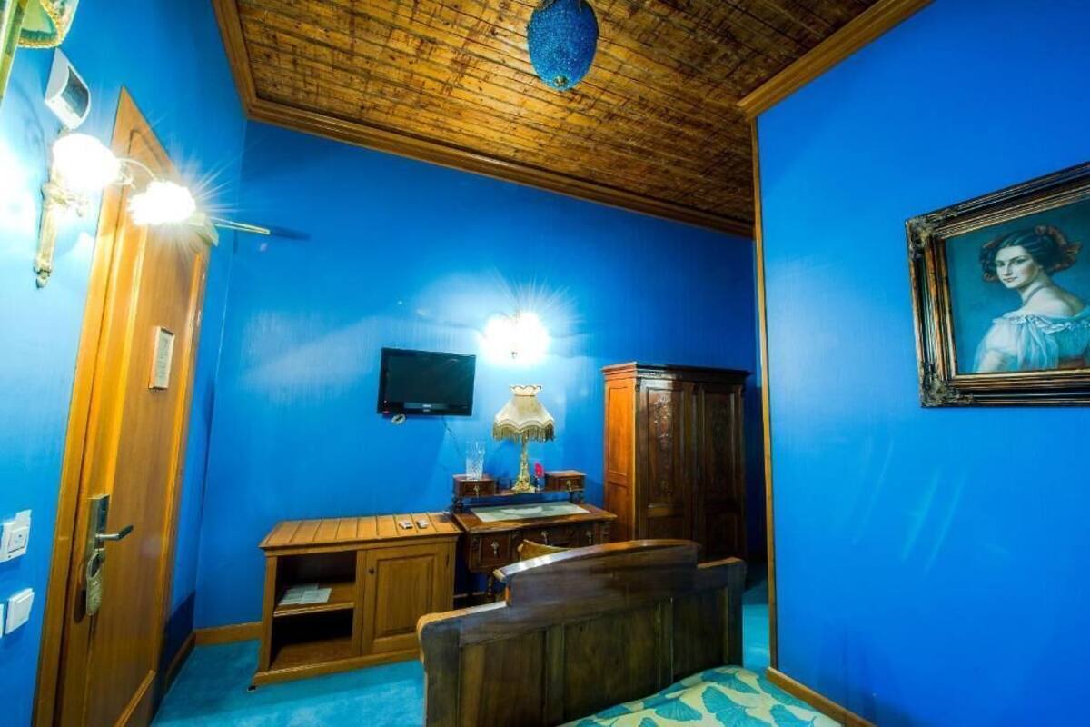 A room with a blue wall, wooden ceiling, a desk with a lamp, a bed, and a framed portrait.