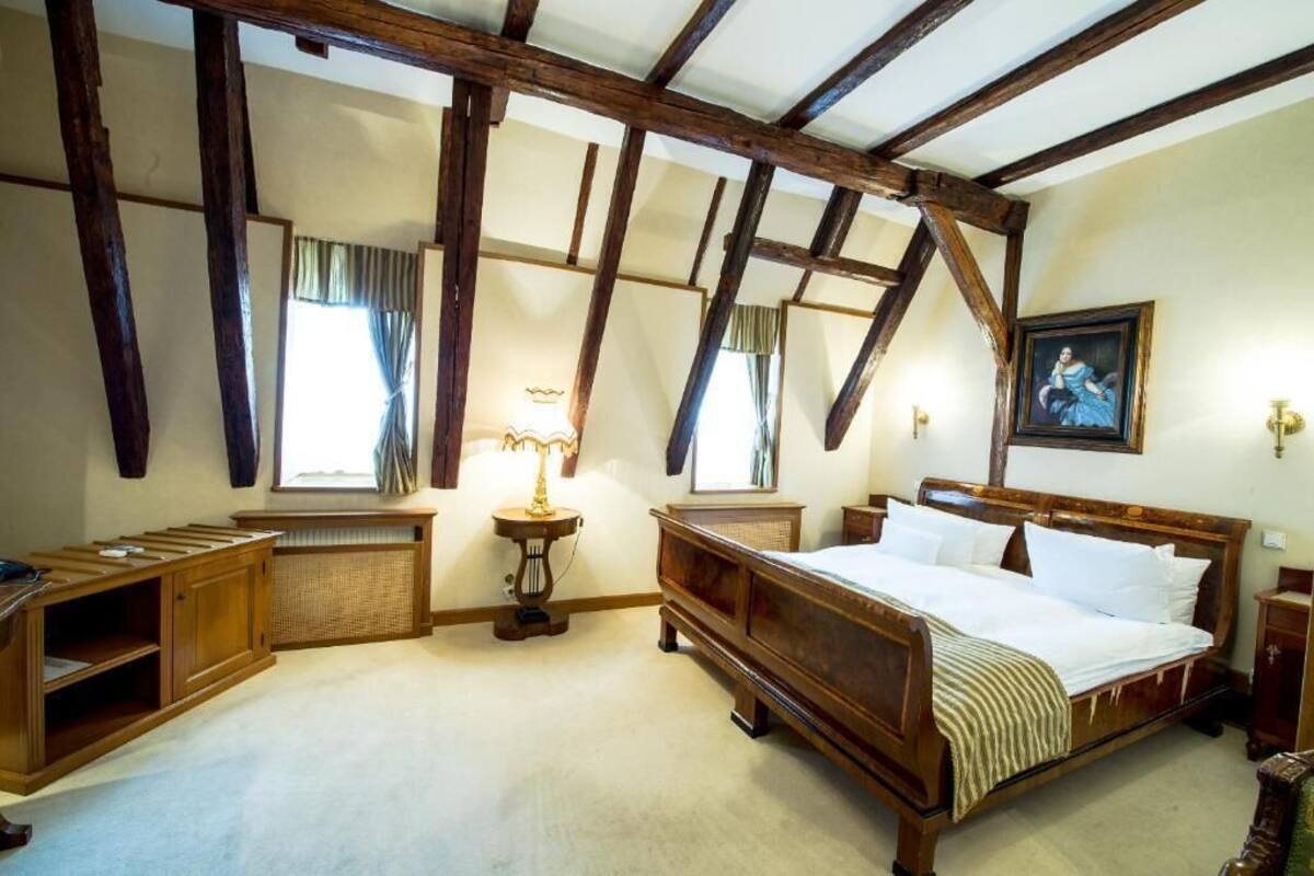 A traditional bedroom with exposed wooden beams, a large bed, a bedside table, a lamp, and a window with curtains.