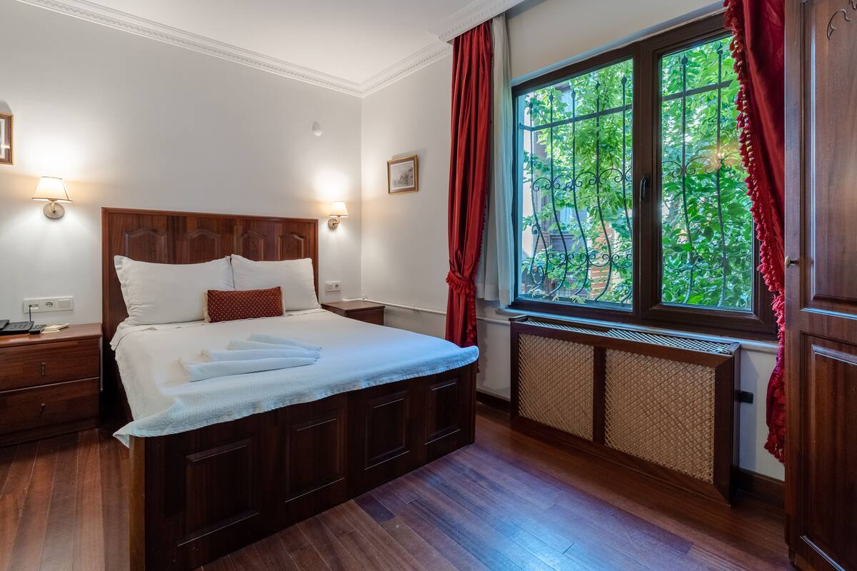A neatly made bed with pillows and a blanket, a bedside table with a lamp, and a window with a view of greenery.
