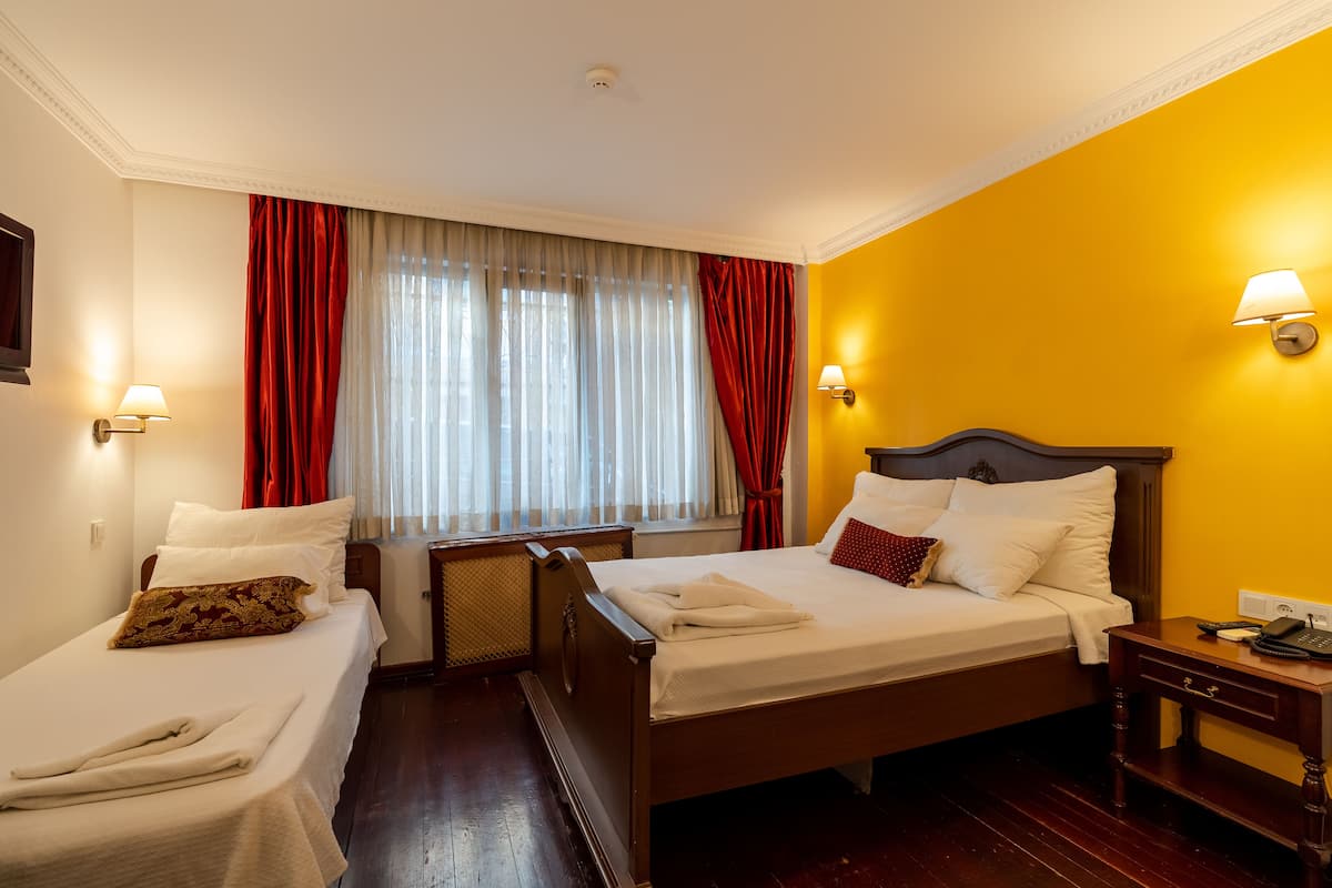 A hotel room with two single beds, a wooden headboard, a nightstand, a lamp, and a window with curtains.