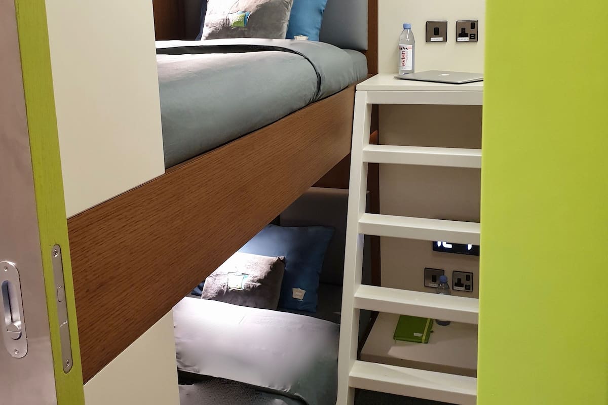 Ein Etagenbett mit Kopfteil, ein Nachttisch mit einer Flasche und Steckdosen an der Wand.