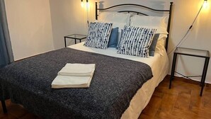 Una cama bien hecha con una manta gris y almohadas, una mesita de noche con una lámpara y un cuadro en la pared.