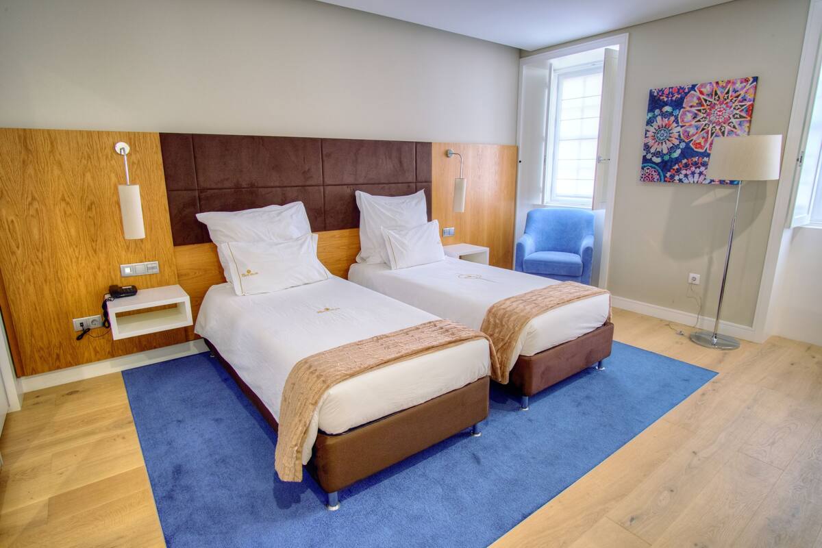 A hotel room with two beds, a blue carpet, wooden flooring, a blue chair, and a colorful wall painting.