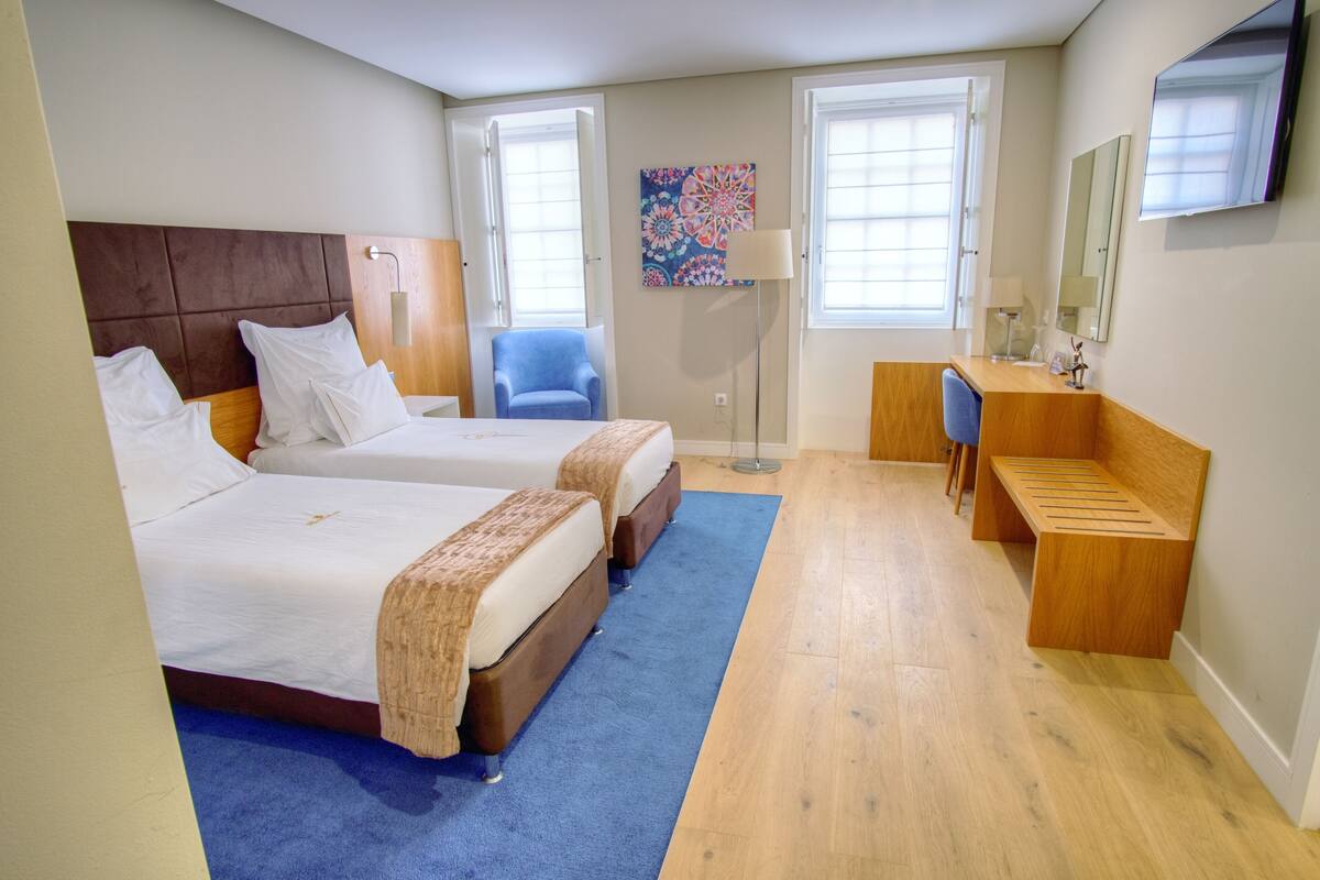 A hotel room with two beds, a desk, a chair, a lamp, and a large window.