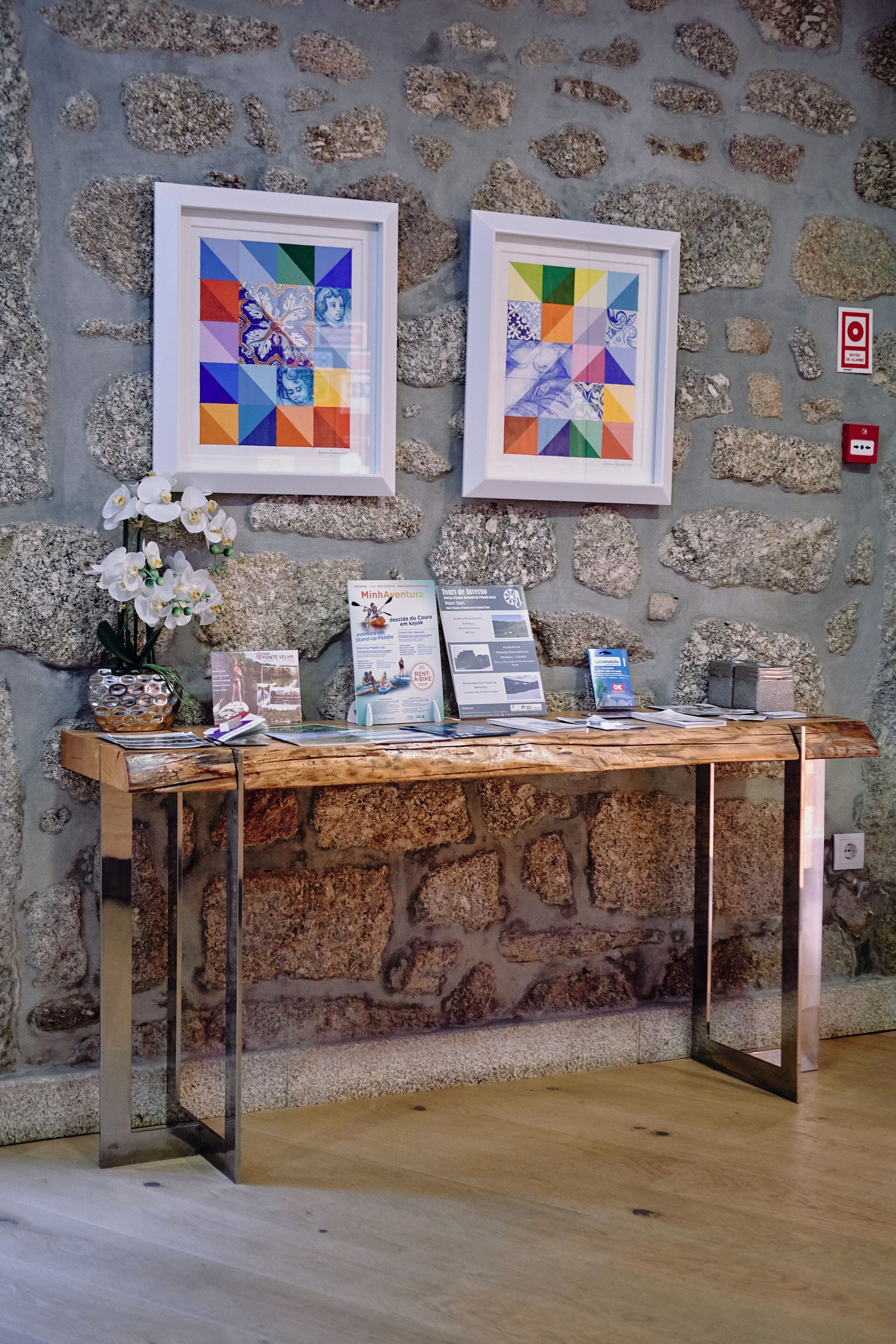 Un mostrador de recepción con obras de arte enmarcadas, folletos informativos y un jarrón con flores blancas frente a una pared de piedra.
