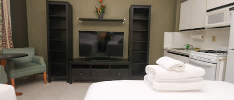 Quarto de hotel com cama, televisão, cozinha compacta e área de estar com mesa e cadeira.