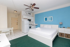 A hotel room with a bed, a desk, a kitchenette, and a ceiling fan.
