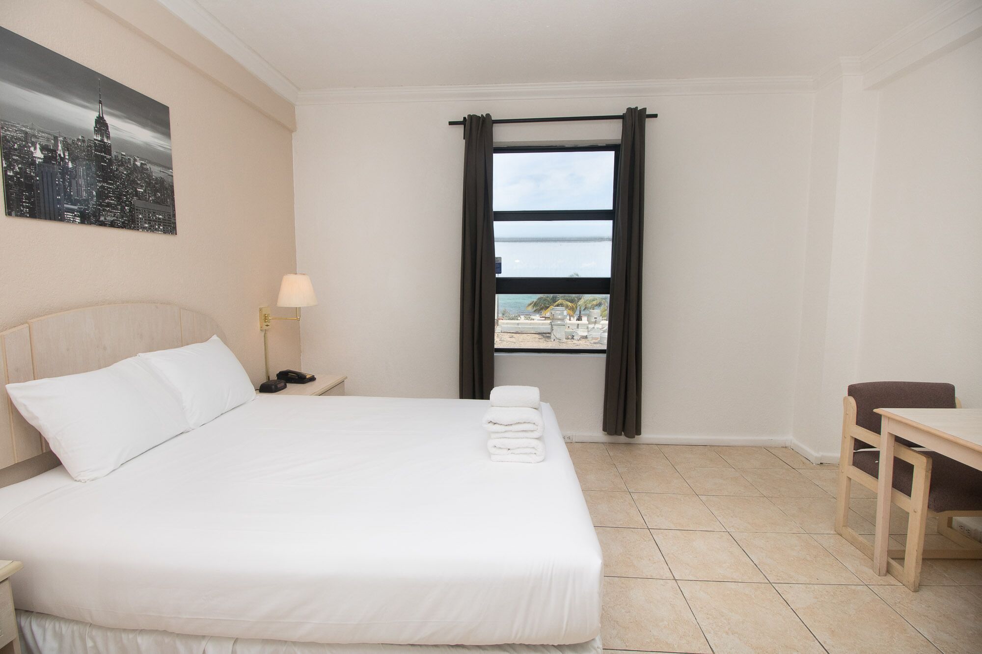 A hotel room with a bed, a nightstand with a lamp, a desk, and a window with curtains.