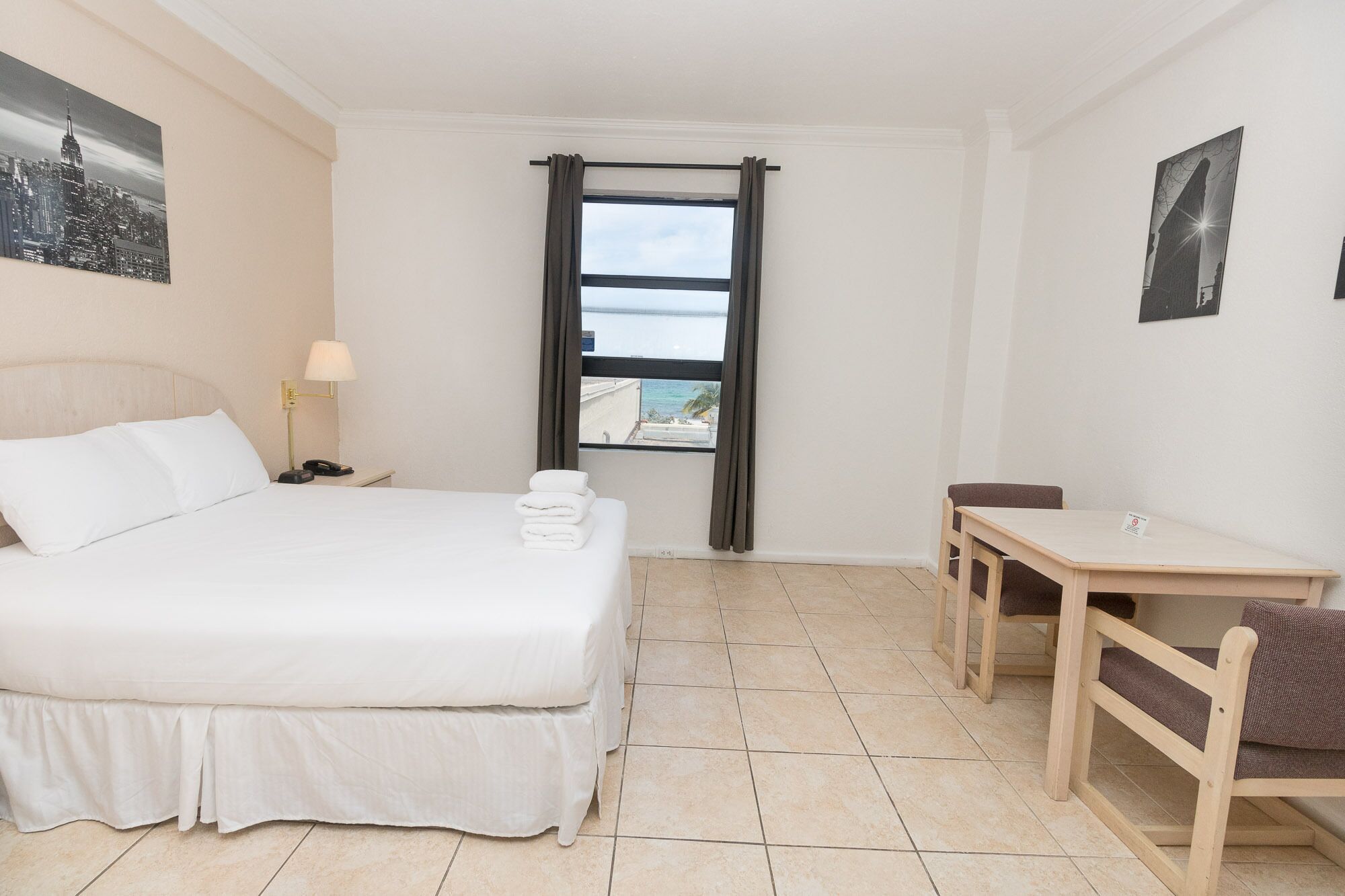 A hotel room with a bed, a desk, a chair, a nightstand with a lamp, and a view of the sea through a window.