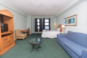 A hotel room with a bed, a blue sofa, a wooden TV stand, a glass coffee table, and a desk with a chair.