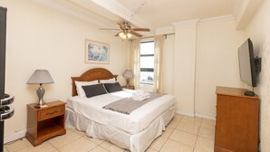 A hotel room with a bed, two bedside lamps, a ceiling fan, a painting, a window with curtains, and a TV on a wooden stand. - 784 Ocean View Hollywood Beach (Hollywood)